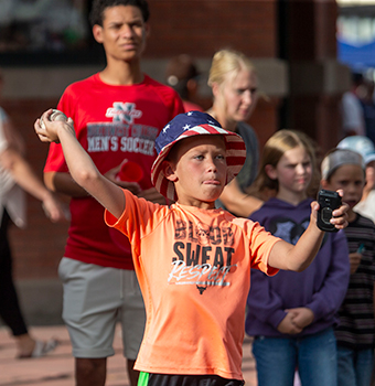 A young boy in an orange shirt and wearing a U.S. flag hat throwing a baseball while others watch