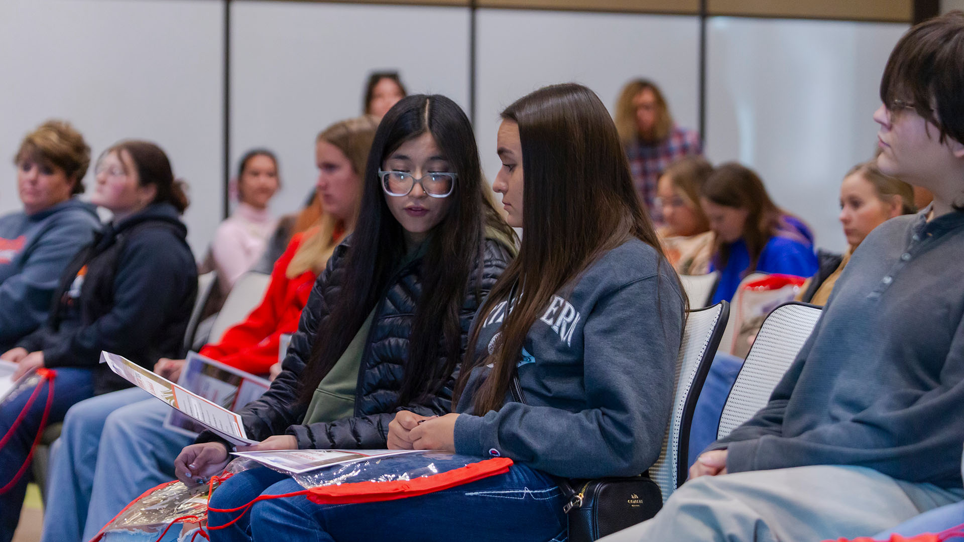 Two female high school students wearing sweaters and jackets looking down at a flyer sitting in a large room surrounded by many other people