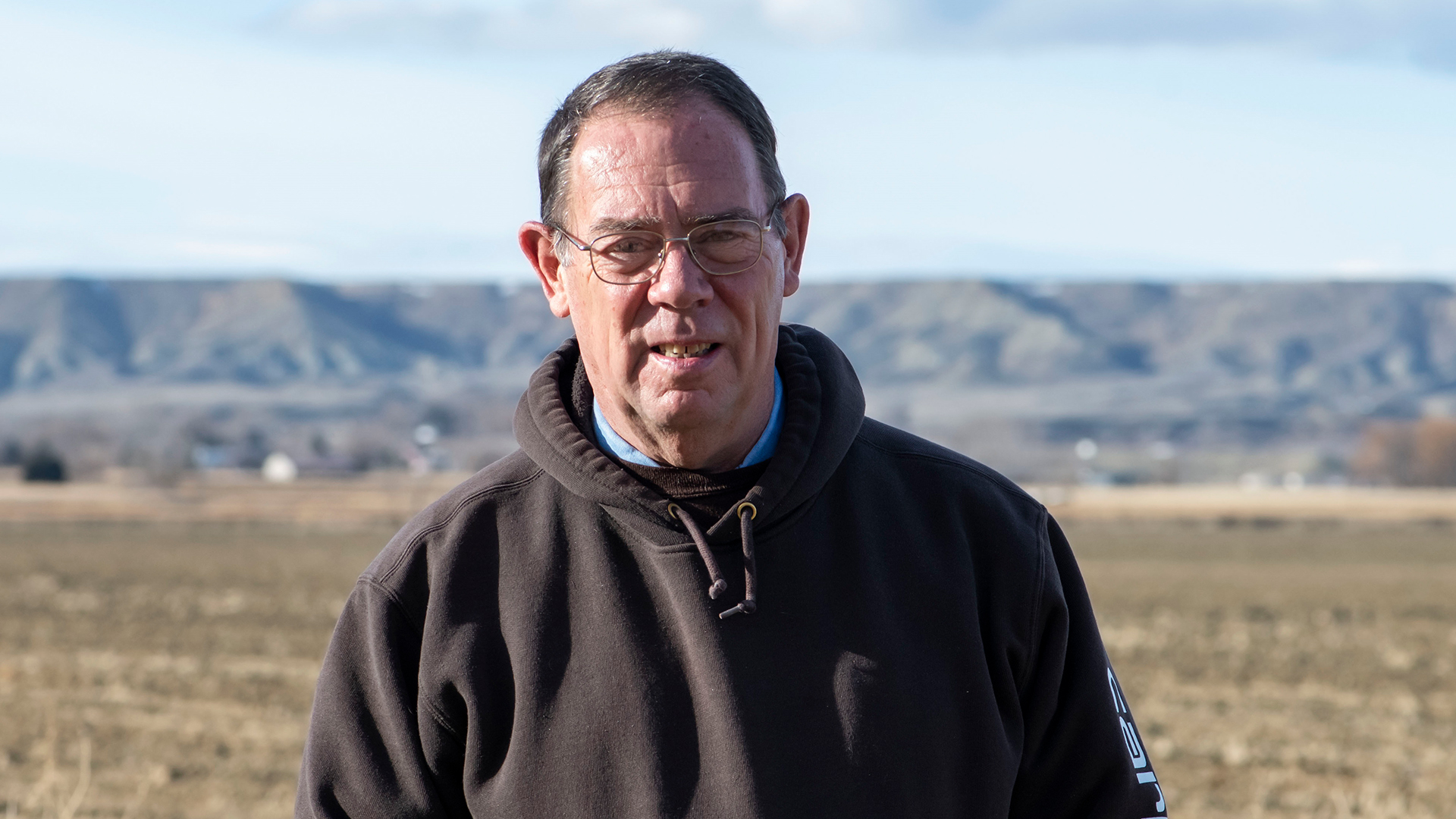 Rob Stothart wearing glasses and standing in a field with low hills behind him