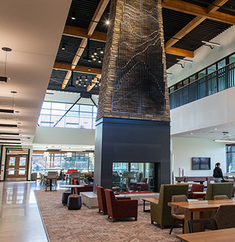 First floor view of floor to ceiling fireplace surrounded by seating area with comfortable chairs and tables and sunlight coming through windows