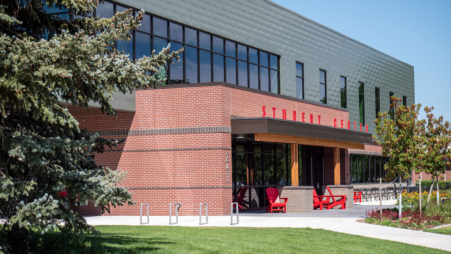 Outside picture of the student center with red brick entryway framing red chairs with a green facade and tall windows on second floor, along with trees in front