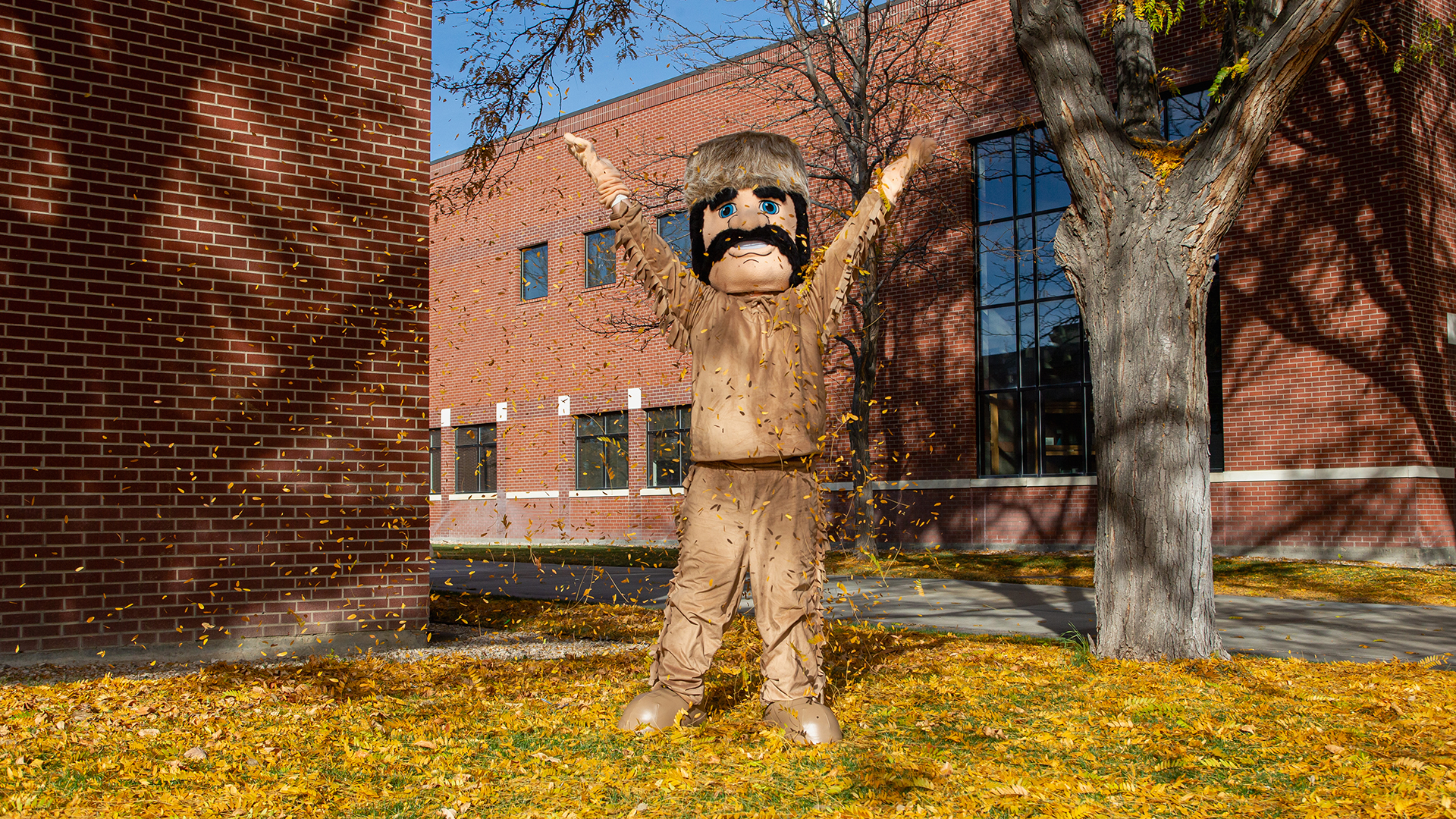 Trapper mascot throwing yellow fall leaves in the air in front of a brick building with large glass window