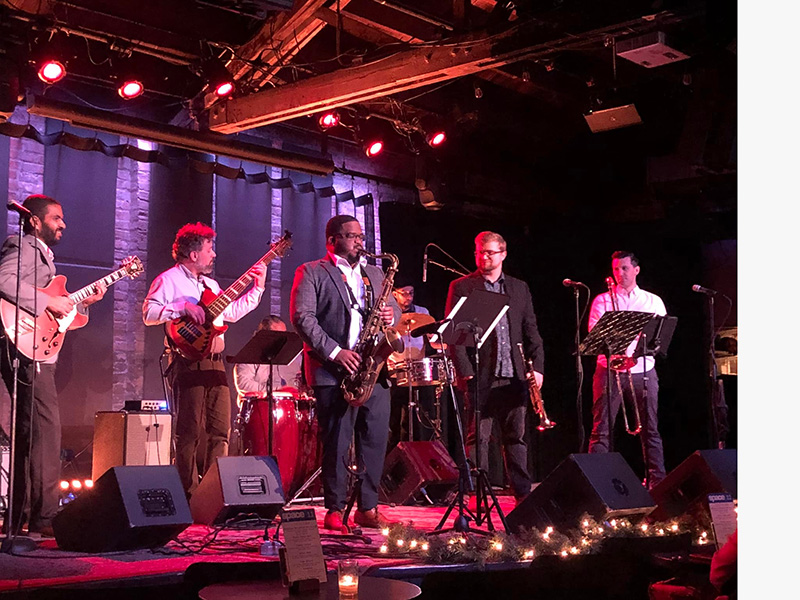 A group of male musicians playing jazz instruments on a stage in front of small candle-lit tables surrounded by speakers and microphones
