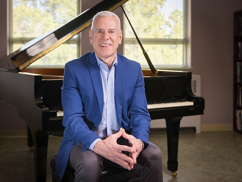 A man with short gray hair sitting in front of a grand piano in a studio setting with a window showing brightly lit trees behind him