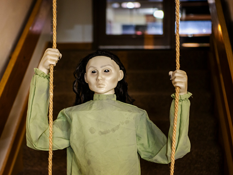 A creepy mannequin in an olive green smock holding onto ropes that support a swing with stairs climbing in the background