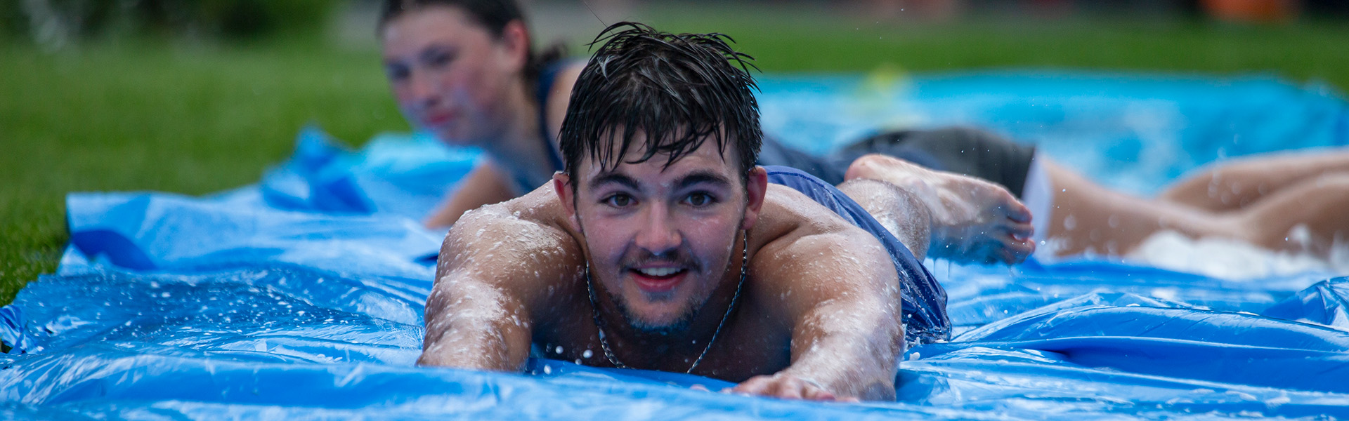 A smiling male student sliding down a blue slip and slide toward the camera