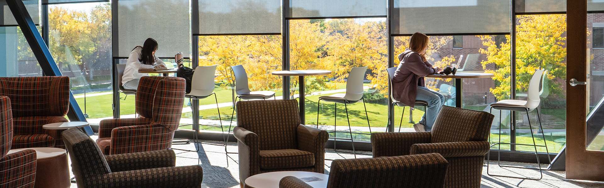 Two students studying at high top tables in front of large second floor windows on a sunny fall day