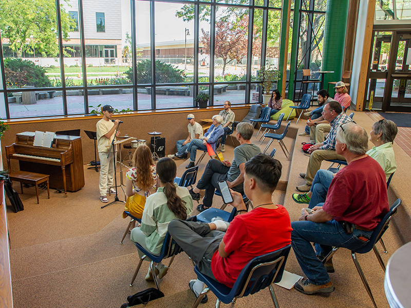 A group of people sitting in a sunken amphitheater listening to a speaker at a podium surrounded by floor to ceiling windows on a sunny day