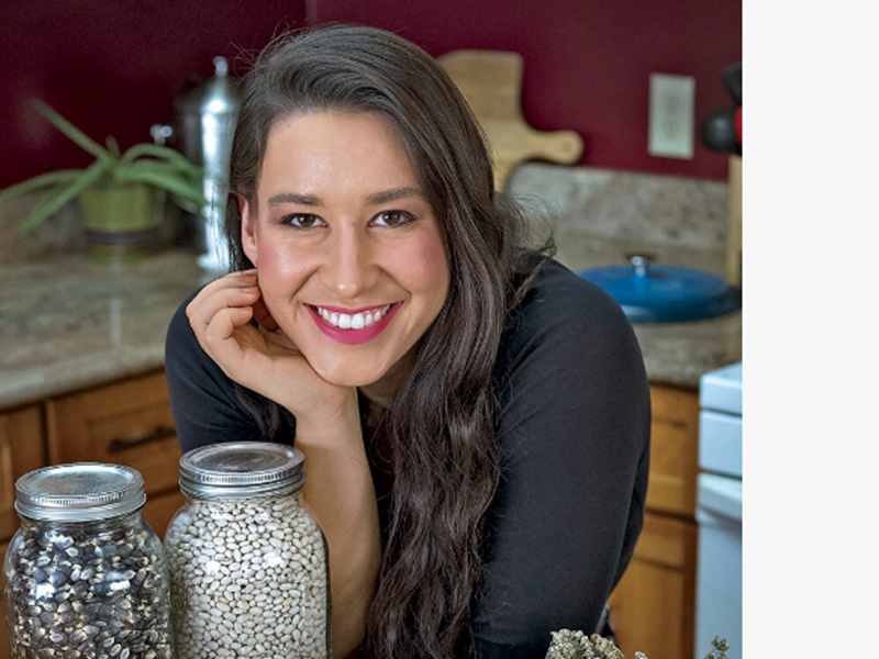 A woman with long dark hair with her chin resting on one hand propped up on a kitchen table, smiling at the camera with blurred kitchen counters behind her