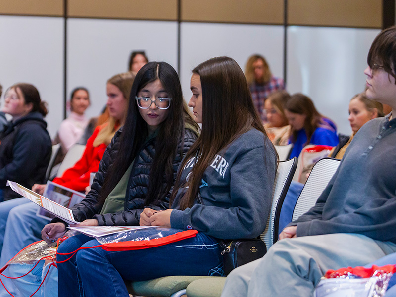 Two female high school students wearing sweaters and jackets looking down at a flyer sitting in a large room surrounded by many other people
