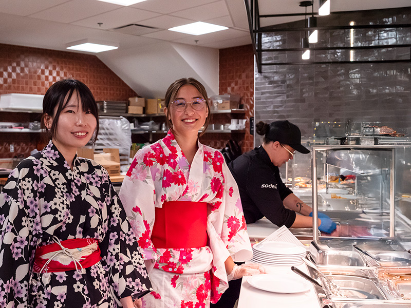 Two smiling young females wearing traditional Japanese clothing standing behind a food station in a cafeteria preparing to serve people