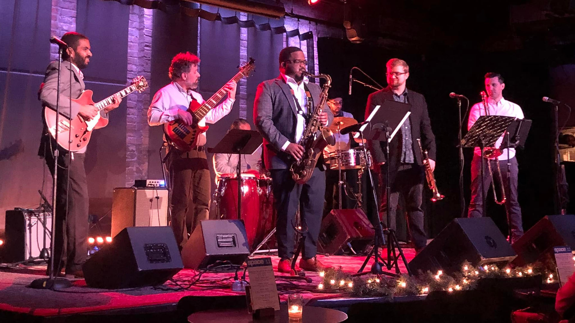 A group of male musicians playing jazz instruments on a stage in front of small candle-lit tables surrounded by speakers and microphones