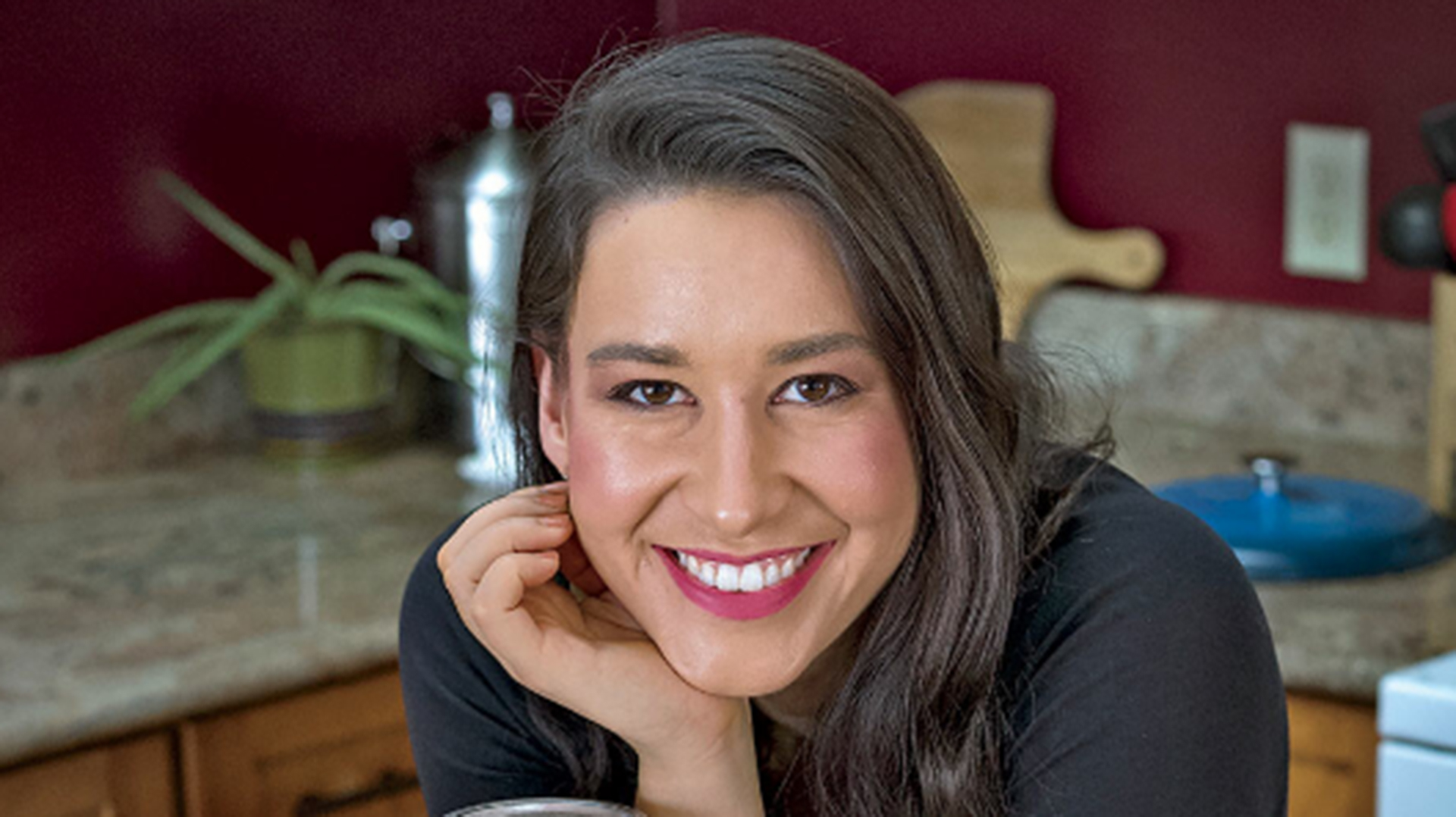 A woman with long dark hair with her chin resting on one hand propped up on a kitchen table, smiling at the camera with blurred kitchen counters behind her