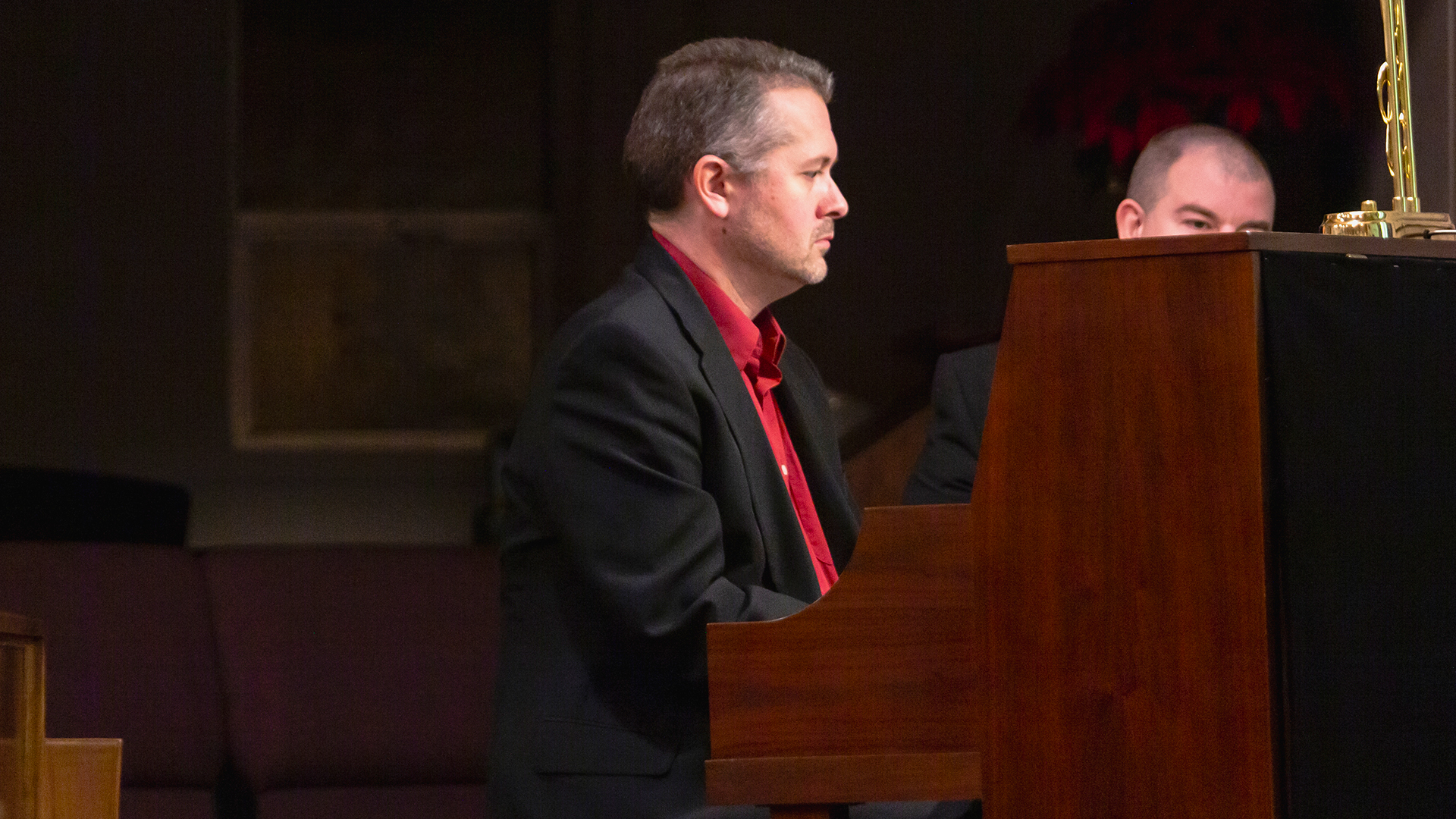 A man in a black coat and red shirt playing a church piano lighted by a lamp on top